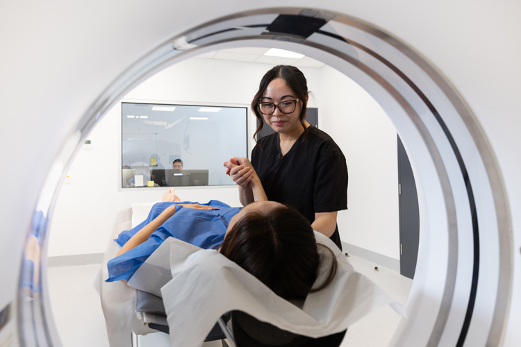 A kind CT radiographer stands beside a patient lying on the scanner table, preparing the patient for a scan.