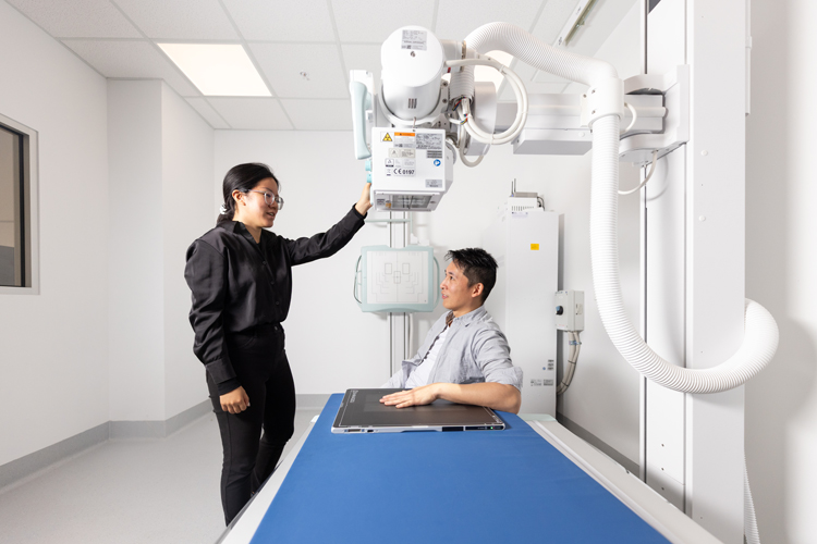 An X‑ray technician positions the X‑ray machine above a patient seated at an examination table.